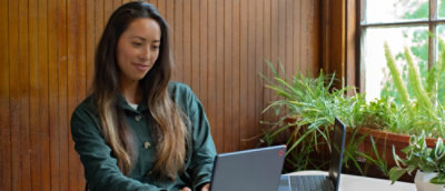 A woman with long hair sitting in front of a laptop with plants in the background.