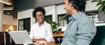 Two individuals seated in an office, discussing while interacting with a laptop.