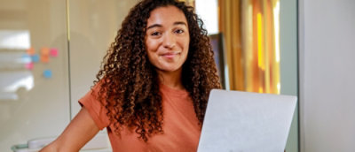 A person with curly hair smiles warmly at the camera while holding a laptop.