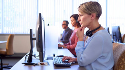 A woman wears headphones and types on a keyboard at a computer station among others in a bright office setting.