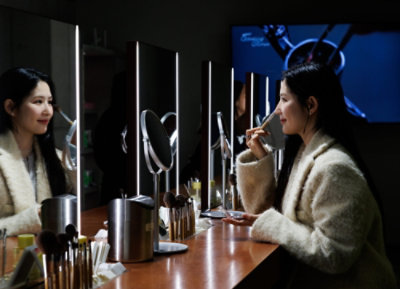 A woman is sitting in front of a mirror, applying makeup to her face with a brush.