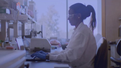 A person in a lab coat and gloves working at a computer.