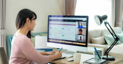 A woman in a pink sweater attends a video conference on a desktop computer in a home office. 