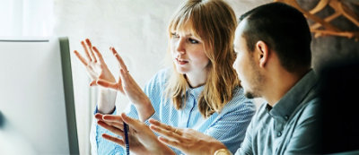 A woman and a man are sitting down and pointing at a computer screen, engaging in a discussion.