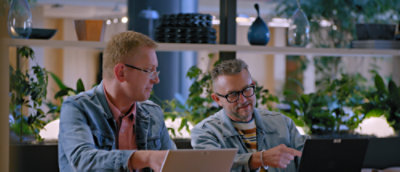 A man wearing glasses and a denim jacket sitting at a table with another man.