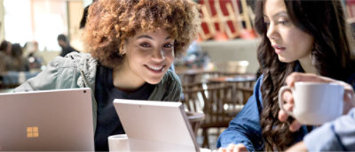 Two people sit at a table in a cafe, each working on a laptop. One person smiles while looking at the other's screen.