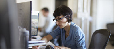 A woman with short hair and glasses works at a computer desk in an office.