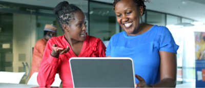 Two women are smiling and looking at a laptop together in an office setting.