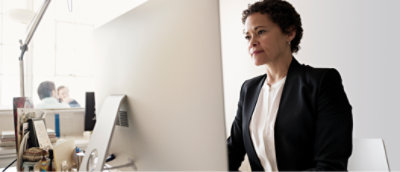 A woman wearing a white shirt and black blazer is seated at a desk, intently working on a desktop computer.