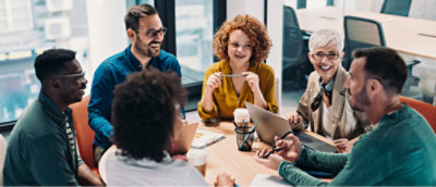A diverse group of six people sits around a table in a modern office, engaged in a discussion.