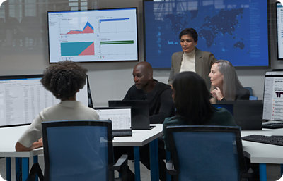 A diverse group of five people are seated around a conference table with multiple computer screens displaying graphs