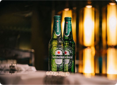 Two Heineken beer bottles on a table with a blurred background of vertical lights.