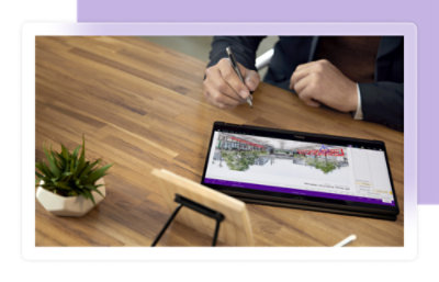 A person writes with a stylus on a tablet displaying an architectural design, next to a small plant on a wooden desk..