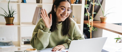 A woman wearing glasses and a green sweater waves at her laptop screen, sitting at a desk with books.