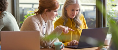 Two women are sitting at a table, looking at a laptop. One is pointing at the screen, and both appear engaged in discussion. 