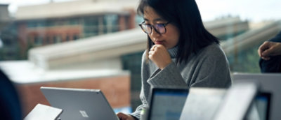 A woman wearing glasses has her hand on her chin and looking down at laptop