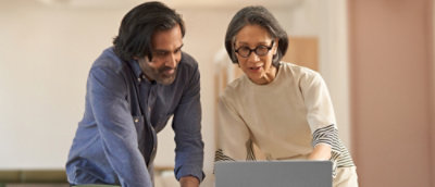 A man with long hair and a beard and a woman in glasses and a white shirt are looking at a laptop.