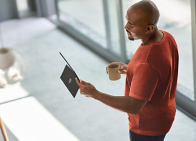 Person standing near a window holding a tablet and a coffee mug while reviewing content in a modern office space.