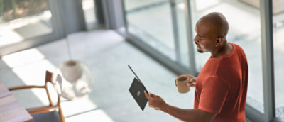 Person standing near a window using a tablet and holding a cup.