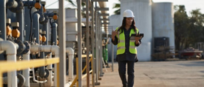 Worker wearing safety gear walking through an industrial facility while using a tablet.