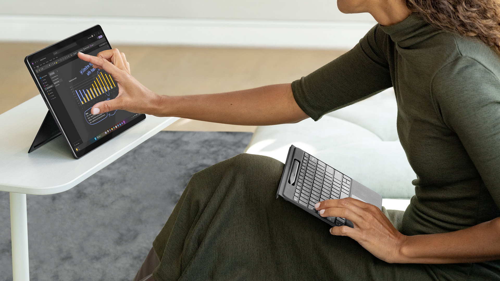 En person bruger et aftaget Surface Pro Flex Keyboard med Slim Pen, til 13 tommer, i skødet, mens vedkommende interagerer med touchskærmen på en Surface-enhed, der er placeret på et bord i nærheden.