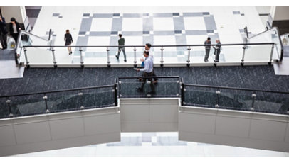 Aerial view of office building‘s interior foyer. Two men are walking across elevated walkway. Man and woman on escalator, various others walking on ground floor below.