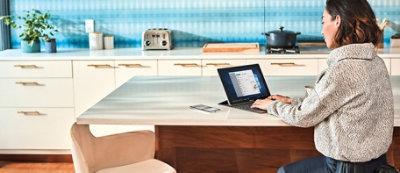 A person sits at a kitchen table using a laptop. The background includes a toaster, a pot on the stove, and other kitchen items.
