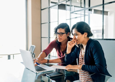 Two women are working together at a glass table, looking at a laptop screen. 