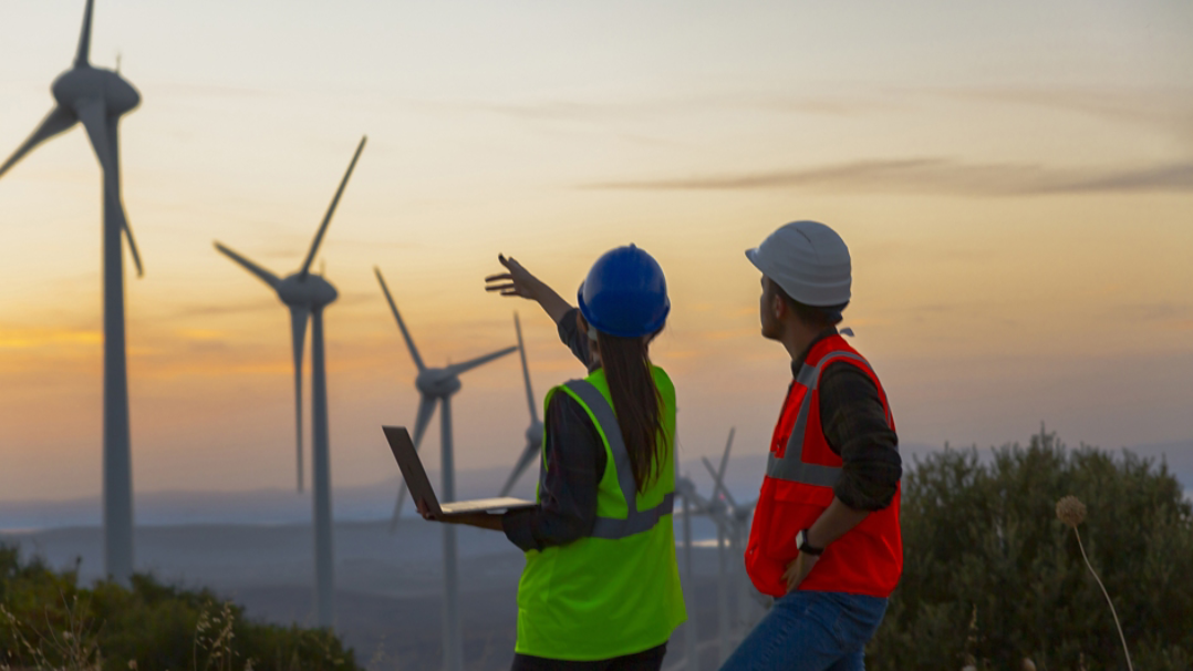 Deux personnes vêtues d’un gilet de chantier regardent un champ d’éoliennes tandis que l’une d’entre elles tient un ordinateur portable