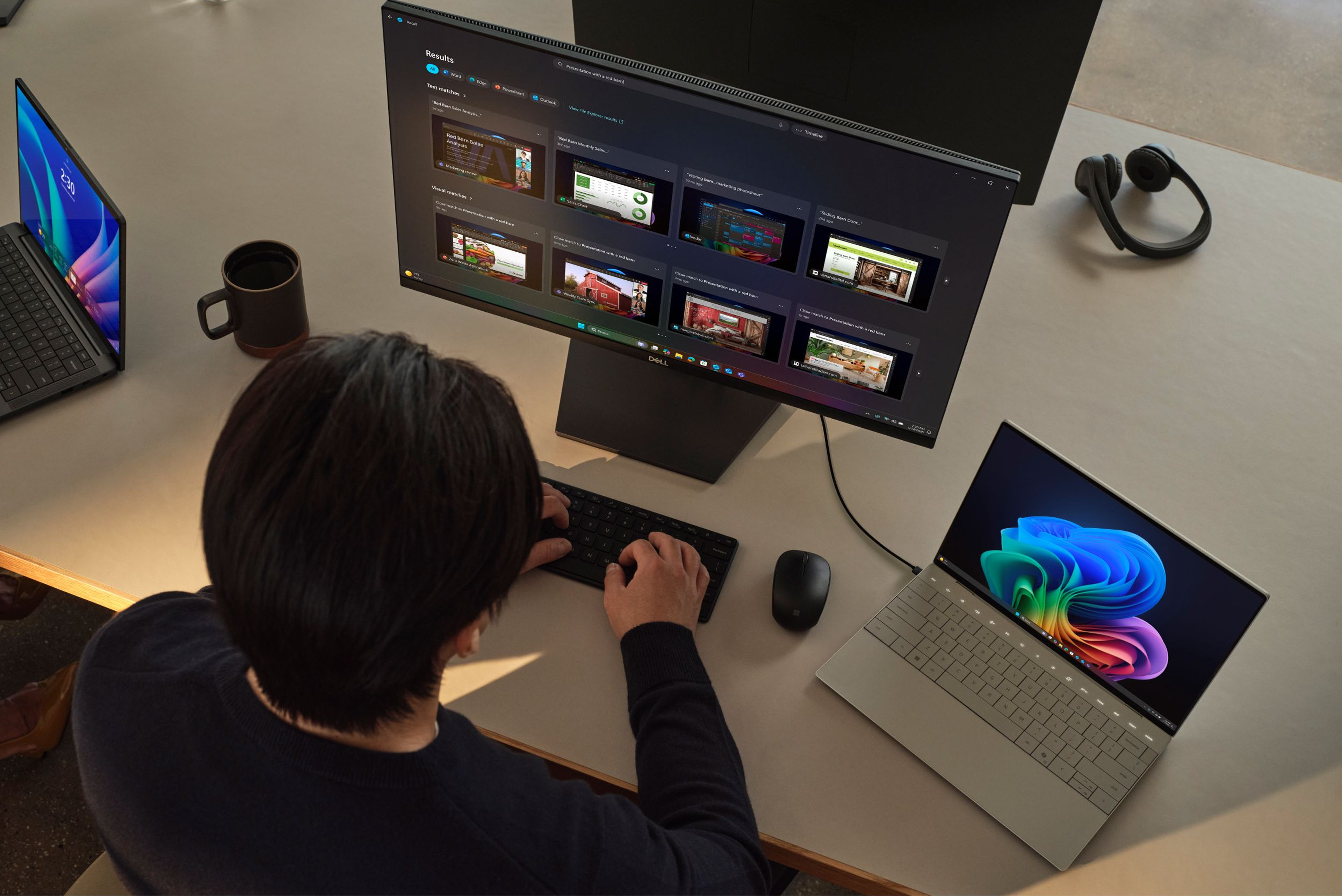 A person sitting at a desk working on a keyboard with a monitor, with two open laptops, a mouse, and headphones