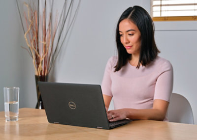 A woman seated at a table works on a Dell laptop with a glass of water next to her.