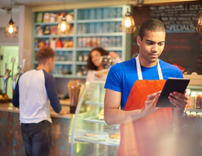 A café worker in an orange apron uses a tablet while another person stands at the counter. 