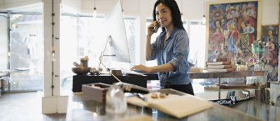 A woman is talking on the phone while working on a computer at a desk in a well-lit room.