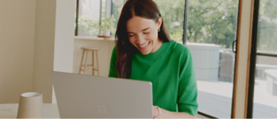 Person in a green sweater smiling while working on a laptop at a table near a window with trees outside.